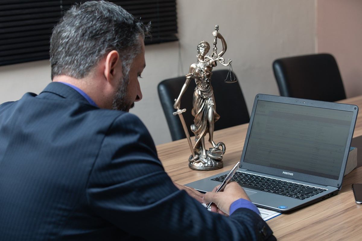 Homme écrivant sur un papier devant un ordinateur portable avec une statue de justice à côté